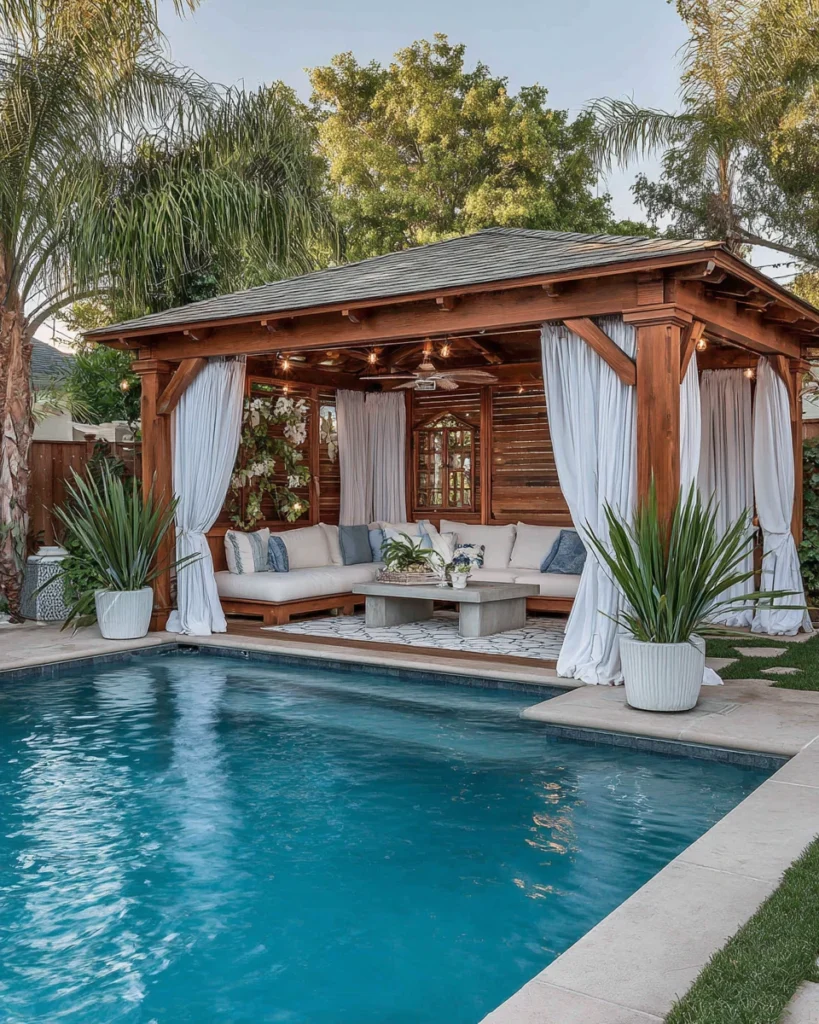 Open-air pool cabana with flowing curtains and outdoor seating beside a backyard swimming pool